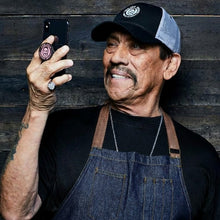 Load image into Gallery viewer, A man wearing a black t-shirt, dark denim apron, and a black-and-gray trucker hat takes a mirror selfie against a dark wood plank wall.
